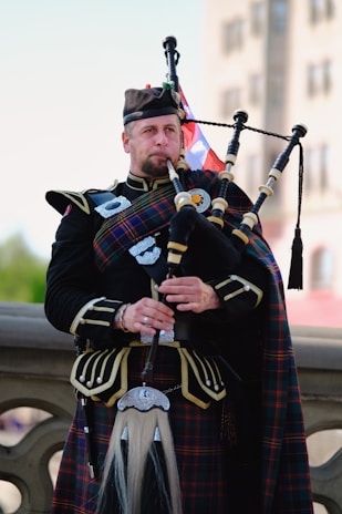 A man dressed in traditional Scottish attire plays a set of bagpipes. He wears a kilt with a tartan pattern and a matching sash, along with a dark jacket adorned with silver buttons. The background shows a blurred outdoor setting with a building and sky visible.