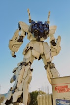 A large, detailed robot stands against a clear blue sky. The robot is predominantly white with black and blue accents, showcasing intricate mechanical features and joints. Its massive size is emphasized by the perspective, making it appear towering and imposing.