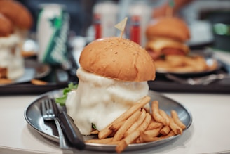 A large burger topped with melted cheese sits on a silver plate, accompanied by a serving of French fries. The burger's sesame seed bun and thick cheese layer are prominent. In the background, other similar meals and drink cans are visible, suggesting a casual dining setting.