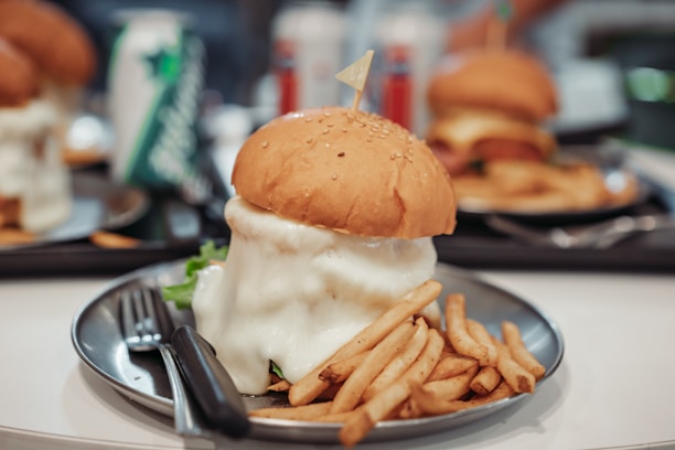 A large burger topped with melted cheese sits on a silver plate, accompanied by a serving of French fries. The burger's sesame seed bun and thick cheese layer are prominent. In the background, other similar meals and drink cans are visible, suggesting a casual dining setting.