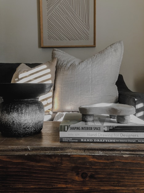 A cozy and minimalist interior setup featuring a stack of design books on a wooden table. There is a textured black vessel and a sculptural object on top of the books. In the background, a large, soft pillow and a light-colored pillow with sunlight creating stripes are on a dark sofa. A framed abstract line art piece hangs on the wall.