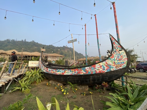 A traditional wooden boat with vibrant, colorful patterns is displayed outdoors. The boat is mounted on a grassy area surrounded by lush green plants and flowers. Several poles with string lights hang overhead. In the background, there are thatched-roof huts and a person walking nearby. The environment suggests a relaxed, rustic outdoor setting.