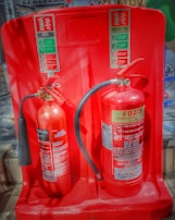 Two fire extinguishers are positioned on a red stand, one labeled as CO2 and the other as Foam. Both extinguishers have instructional labels with icons and text, indicating their uses and safety precautions.