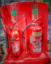 Two fire extinguishers are positioned on a red stand, one labeled as CO2 and the other as Foam. Both extinguishers have instructional labels with icons and text, indicating their uses and safety precautions.