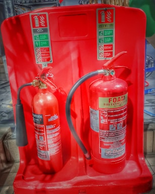Two fire extinguishers are positioned on a red stand, one labeled as CO2 and the other as Foam. Both extinguishers have instructional labels with icons and text, indicating their uses and safety precautions.