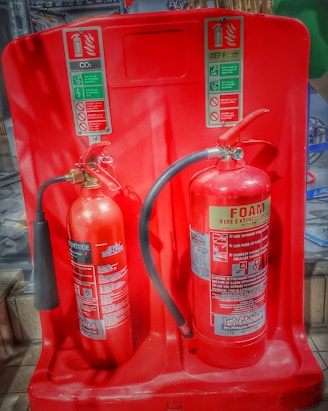 Two fire extinguishers are positioned on a red stand, one labeled as CO2 and the other as Foam. Both extinguishers have instructional labels with icons and text, indicating their uses and safety precautions.