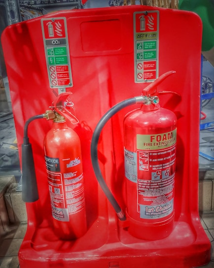 Two fire extinguishers are positioned on a red stand, one labeled as CO2 and the other as Foam. Both extinguishers have instructional labels with icons and text, indicating their uses and safety precautions.