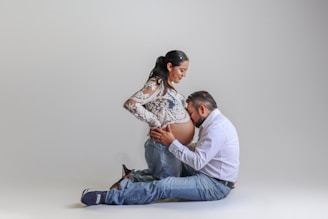 A pregnant woman stands with a gentle smile while a man kneels in front of her, kissing her belly. Both are dressed casually, with the woman wearing a sheer lace top and jeans, and the man in a white shirt and jeans. The background is a simple, soft gradient, emphasizing their connection.