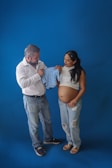 A couple stands together against a blue background, both looking at a small blue baby onesie. The woman is pregnant, wearing a white top and ripped jeans, while the man wears a white shirt and jeans. They appear focused and content as they discuss the baby clothing.