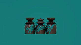 Three sculptural objects with a glossy brown finish are aligned side by side. They are decorated with bold, blue graffiti-style text. Each object has a unique shape, with the middle one being more intricately designed. The background is a solid turquoise color.