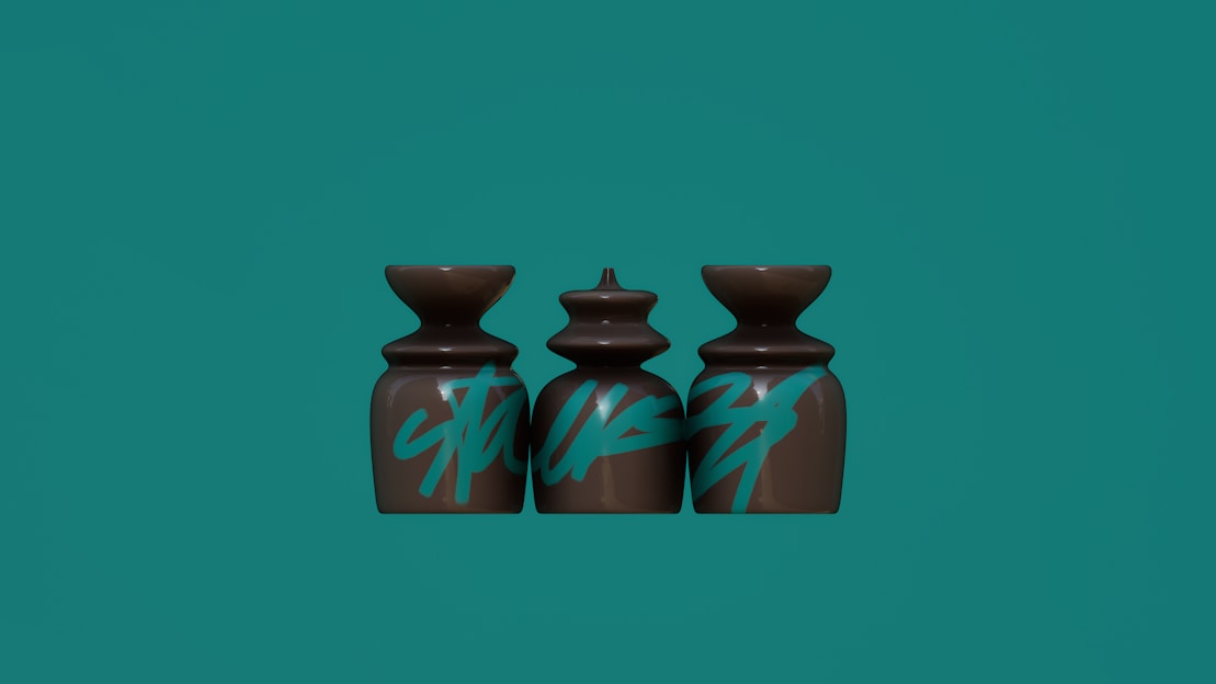 Three sculptural objects with a glossy brown finish are aligned side by side. They are decorated with bold, blue graffiti-style text. Each object has a unique shape, with the middle one being more intricately designed. The background is a solid turquoise color.