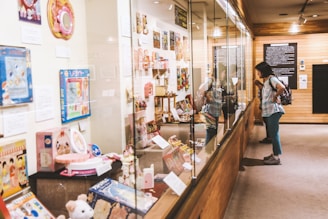 A person is closely observing a display case filled with a variety of vintage toys and promotional materials in a well-lit museum setting. Posters and information plaques are mounted on the walls alongside the collection of nostalgic items.