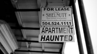 A black and white photograph of a real estate sign hanging from a building. The sign reads 'FOR LEASE' with the contact information for SHELNUTT Real Estate Enterprises. Beneath the main sign, there is an additional sign that says 'APARTMENT HAUNTED'. The angle of the photo shows the underside of a roof and support structure.