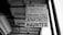 A black and white photograph of a real estate sign hanging from a building. The sign reads 'FOR LEASE' with the contact information for SHELNUTT Real Estate Enterprises. Beneath the main sign, there is an additional sign that says 'APARTMENT HAUNTED'. The angle of the photo shows the underside of a roof and support structure.