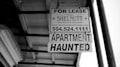 A black and white photograph of a real estate sign hanging from a building. The sign reads 'FOR LEASE' with the contact information for SHELNUTT Real Estate Enterprises. Beneath the main sign, there is an additional sign that says 'APARTMENT HAUNTED'. The angle of the photo shows the underside of a roof and support structure.
