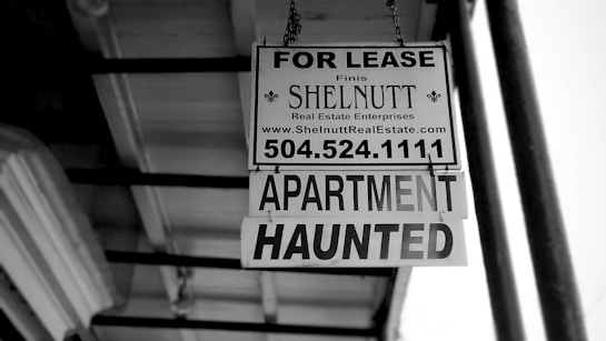A black and white photograph of a real estate sign hanging from a building. The sign reads 'FOR LEASE' with the contact information for SHELNUTT Real Estate Enterprises. Beneath the main sign, there is an additional sign that says 'APARTMENT HAUNTED'. The angle of the photo shows the underside of a roof and support structure.