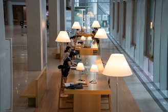 A modern interior space with long wooden tables arranged in a row, each illuminated by large, stylish lamps. People are seated at the tables, engaged in activities such as working on laptops and reading documents. The space is characterized by high ceilings, tall windows, and a sleek, minimalist design.