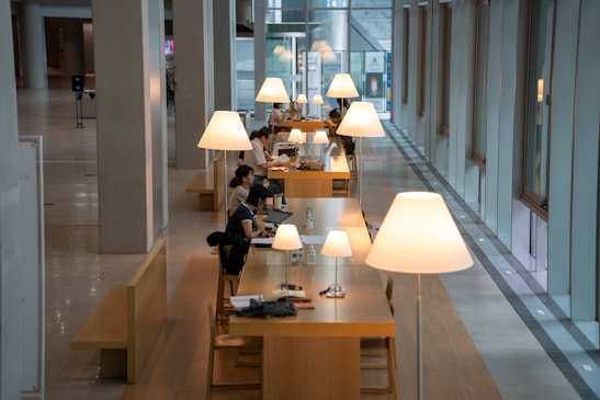 A modern interior space with long wooden tables arranged in a row, each illuminated by large, stylish lamps. People are seated at the tables, engaged in activities such as working on laptops and reading documents. The space is characterized by high ceilings, tall windows, and a sleek, minimalist design.