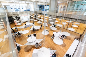 A spacious, modern interior with a wooden floor, filled with round tables and multiple people sitting around them engaged in various activities. Circular light fixtures hang from the high ceiling, creating an open and airy feel. Large windows and a glass railing provide a sense of openness and contemporary design.
