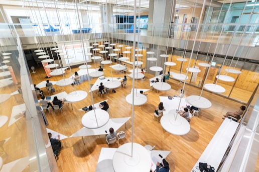 A spacious, modern interior with a wooden floor, filled with round tables and multiple people sitting around them engaged in various activities. Circular light fixtures hang from the high ceiling, creating an open and airy feel. Large windows and a glass railing provide a sense of openness and contemporary design.