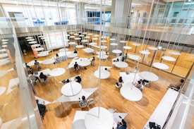 A spacious, modern interior with a wooden floor, filled with round tables and multiple people sitting around them engaged in various activities. Circular light fixtures hang from the high ceiling, creating an open and airy feel. Large windows and a glass railing provide a sense of openness and contemporary design.