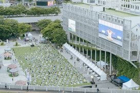 An outdoor event with numerous people practicing yoga on a green field, arranged in neat rows. A large screen on a building displays an instructor demonstrating yoga poses. Several white tents line the perimeter, and there are trees and urban structures in the background.