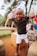 A child with a playful expression is posing outdoors. The child is wearing a camouflage shirt and white shorts. In the background, there are other children playing, along with greenery and trees suggesting a park environment.
