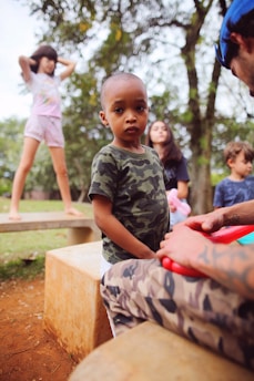 Several children are gathered in an outdoor setting surrounded by trees. One child in a camouflage shirt stands at the center, facing the camera with a serious expression. Other children are engaged in different activities, with one standing on a concrete bench and another looking to the side. An adult with tattoos is partially visible, interacting with the child in the foreground.