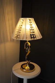 A table lamp with a decorative, transparent shade adorned with crystal-like ornaments. The lamp has a golden, curved metallic stand and is placed on a round white table against a two-toned wall.