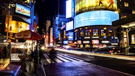 A bustling city street at night with vibrant neon lights and billboards. Vehicles create light trails as they move along the crosswalk, surrounded by illuminated advertisements and a lively urban atmosphere. A food cart with umbrellas is visible in the foreground, adding to the busy city setting.