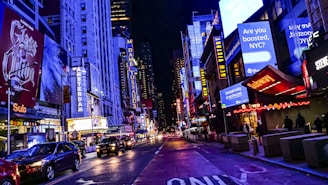 A vibrant city street at night with numerous colorful illuminated billboards and advertisements lining the buildings. The street is bustling with traffic, including cars and pedestrians. Notable signs include those for theaters, restaurants, and vaccination promotions.