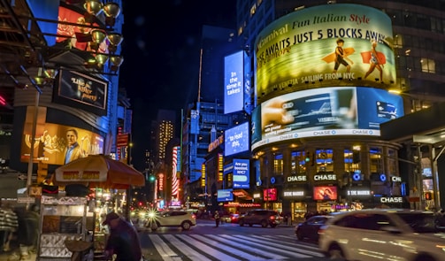 An urban scene bustling with activity at night. Bright digital billboards display advertisements above city streets lined with cars and a hot dog stand. Neon lights and colorful signage create a vibrant atmosphere as pedestrians walk along the sidewalks.