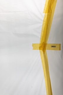 A white surface partially covered with strips of yellow masking tape arranged in an abstract pattern. There is a visible barcode on one piece of tape.