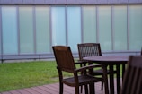 A wooden outdoor patio set consists of a round table and several chairs on a deck. In the background, there is a strip of green lawn and a tall building with large, frosted glass windows.