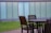A wooden outdoor patio set consists of a round table and several chairs on a deck. In the background, there is a strip of green lawn and a tall building with large, frosted glass windows.