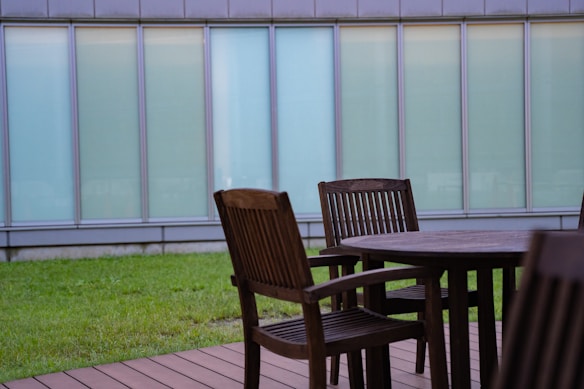 A wooden outdoor patio set consists of a round table and several chairs on a deck. In the background, there is a strip of green lawn and a tall building with large, frosted glass windows.