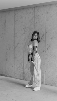 A person stands confidently against a textured wall. They are wearing high-waisted jeans, a crop top, and sneakers. Sunglasses rest on their head, and their gaze is directed to the side. The image is in black and white, emphasizing the contrast and textures.