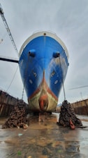 A large blue ship is stationed in a dry dock, resting on a platform. Massive chains are attached to the front, lying in heavy piles on the wet dock floor. The surrounding dock has a weathered and industrial appearance, with a cloudy gray sky overhead.