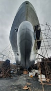 A large ship is positioned on land, surrounded by scaffolding and industrial materials. The bottom view highlights the ship's enormous hull, reflecting a silvery sheen against the overcast sky. Chains and construction equipment are scattered around, indicating an ongoing maintenance or construction process.