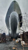 A large ship is positioned on land, surrounded by scaffolding and industrial materials. The bottom view highlights the ship's enormous hull, reflecting a silvery sheen against the overcast sky. Chains and construction equipment are scattered around, indicating an ongoing maintenance or construction process.