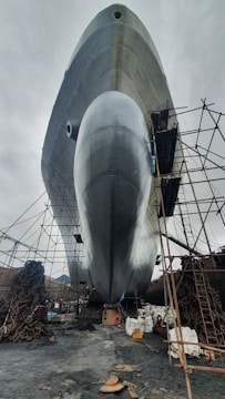 A large ship is positioned on land, surrounded by scaffolding and industrial materials. The bottom view highlights the ship's enormous hull, reflecting a silvery sheen against the overcast sky. Chains and construction equipment are scattered around, indicating an ongoing maintenance or construction process.