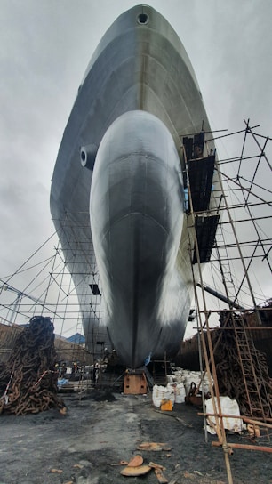 A large ship is positioned on land, surrounded by scaffolding and industrial materials. The bottom view highlights the ship's enormous hull, reflecting a silvery sheen against the overcast sky. Chains and construction equipment are scattered around, indicating an ongoing maintenance or construction process.