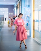 A young woman stands confidently in a shopping mall corridor, wearing a matching pink top and skirt with the word 'Barbie' on the shirt. She is holding a pair of sunglasses in her hand. The background features a modern shopping environment with glass windows showcasing retail stores and a few people visible in the distance.
