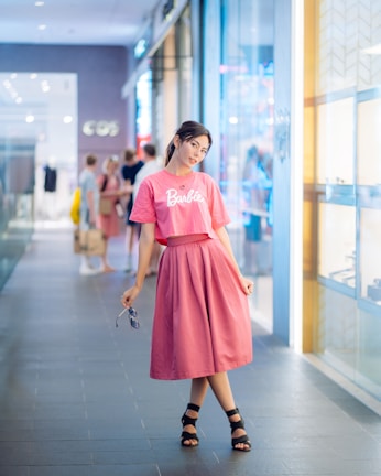 A young woman stands confidently in a shopping mall corridor, wearing a matching pink top and skirt with the word 'Barbie' on the shirt. She is holding a pair of sunglasses in her hand. The background features a modern shopping environment with glass windows showcasing retail stores and a few people visible in the distance.