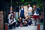 A group of seven young women are gathered together outdoors, posing confidently. They are dressed in a variety of schoolgirl-inspired outfits with plaid skirts, blouses, and some casual wear. The setting appears to be an alleyway or courtyard with a mixture of urban and natural elements. Some are standing while others are sitting or kneeling, giving the scene a dynamic feel.