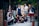 A group of seven young women are gathered together outdoors, posing confidently. They are dressed in a variety of schoolgirl-inspired outfits with plaid skirts, blouses, and some casual wear. The setting appears to be an alleyway or courtyard with a mixture of urban and natural elements. Some are standing while others are sitting or kneeling, giving the scene a dynamic feel.