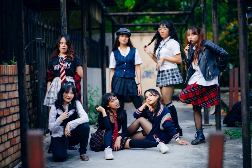 A group of seven young women are gathered together outdoors, posing confidently. They are dressed in a variety of schoolgirl-inspired outfits with plaid skirts, blouses, and some casual wear. The setting appears to be an alleyway or courtyard with a mixture of urban and natural elements. Some are standing while others are sitting or kneeling, giving the scene a dynamic feel.
