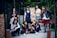 A group of seven young women are gathered together outdoors, posing confidently. They are dressed in a variety of schoolgirl-inspired outfits with plaid skirts, blouses, and some casual wear. The setting appears to be an alleyway or courtyard with a mixture of urban and natural elements. Some are standing while others are sitting or kneeling, giving the scene a dynamic feel.