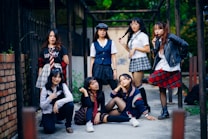 A group of seven young women are gathered together outdoors, posing confidently. They are dressed in a variety of schoolgirl-inspired outfits with plaid skirts, blouses, and some casual wear. The setting appears to be an alleyway or courtyard with a mixture of urban and natural elements. Some are standing while others are sitting or kneeling, giving the scene a dynamic feel.