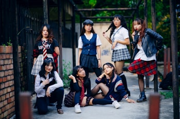 A group of seven young women are gathered together outdoors, posing confidently. They are dressed in a variety of schoolgirl-inspired outfits with plaid skirts, blouses, and some casual wear. The setting appears to be an alleyway or courtyard with a mixture of urban and natural elements. Some are standing while others are sitting or kneeling, giving the scene a dynamic feel.